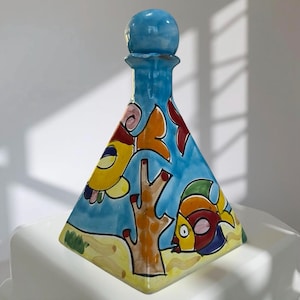 May include: A hand-painted ceramic decanter with a blue body and a round stopper. The decanter features colorful fish and coral designs on a light blue background. The base is yellow, and the overall shape is triangular.