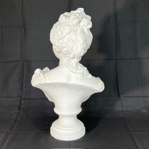 May include: White plaster bust sculpture of a woman with her hair styled in an updo. The bust is on a pedestal base.