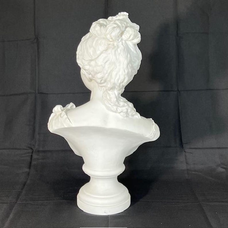 May include: White plaster bust sculpture of a woman with her hair styled in an updo. The bust is on a pedestal base.