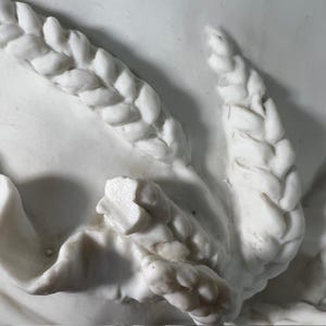 May include: Close-up of a white marble sculpture with intricate braided details. The sculpture features a series of interwoven strands, creating a textured and decorative design.