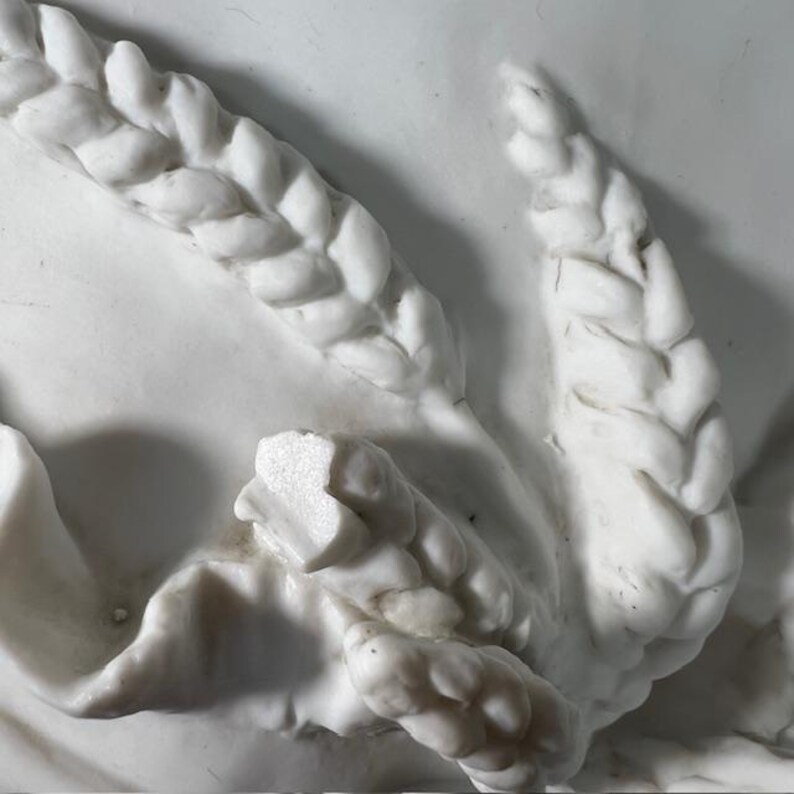 May include: Close-up of a white marble sculpture with intricate braided details. The sculpture features a series of interwoven strands, creating a textured and decorative design.