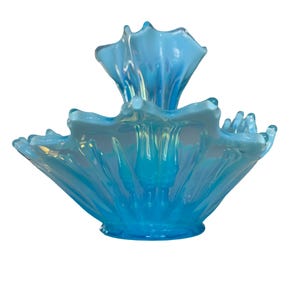 Puede incluir: Un cuenco de vidrio azul claro opalino con un dise&ntilde;o ondulado en forma de p&eacute;talo. El cuenco tiene una base ancha y acampanada y un elemento central curvado hacia arriba. El vidrio tiene un acabado brillante, que refleja la luz. El cuenco tiene aproximadamente 20 cm de di&aacute;metro.