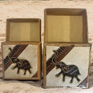 May include: Two wooden boxes with open tops. The boxes have a square shape with a decorative elephant design on the front. The elephant is dark brown and the background is cream-colored with brown diagonal stripes. The boxes are sitting on a sandy surface.