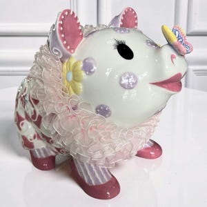 May include: A decorative ceramic piggy bank in the shape of a pig. The pig is white with pink and purple accents, including polka dots and a ruffled collar. A butterfly rests on its snout. The piggy bank has pink shoes.