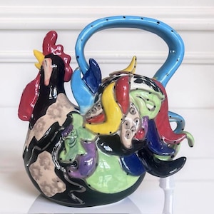 May include: A ceramic teapot shaped like a rooster with a glossy finish. The body features a vibrant, multi-colored abstract pattern in shades of black, red, yellow, green, purple, and blue. It includes a large, arched blue handle with black dots.