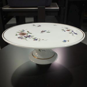 May include: White ceramic cake stand with floral design. The stand features a round top with a gold trim and a pedestal base. The floral design includes purple, blue, and brown flowers and green leaves.