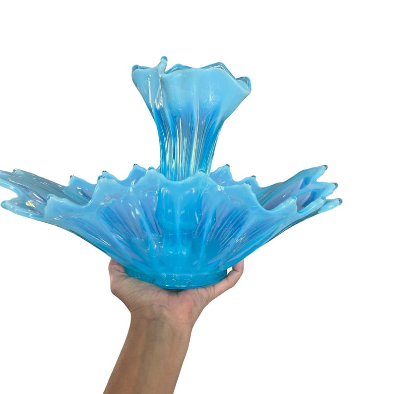 Puede incluir: Un centro de mesa de vidrio azul claro de gran tama&ntilde;o con un dise&ntilde;o &uacute;nico y en capas. La pieza presenta una base ancha y con volantes y un elemento central ascendente, creando una forma art&iacute;stica llamativa. El vidrio tiene una sutil calidad iridiscente.