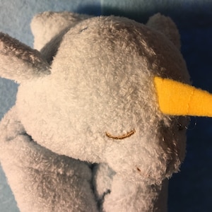 May include: A light blue plush narwhal toy with a yellow horn and embroidered features. The toy has a soft, textured surface and closed eyes, giving a sleepy appearance. The background is a darker blue.