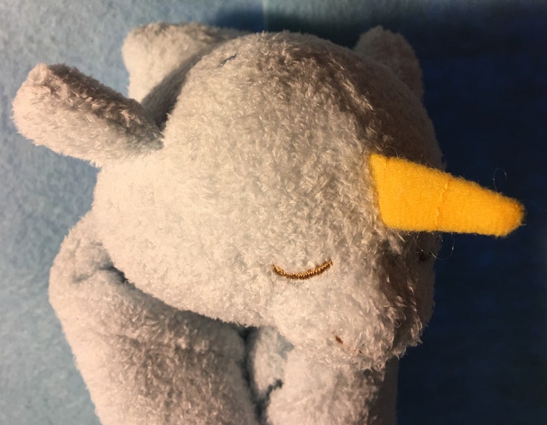 May include: A light blue plush narwhal toy with a yellow horn and embroidered features. The toy has a soft, textured surface and closed eyes, giving a sleepy appearance. The background is a darker blue.