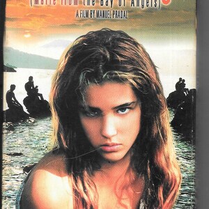 Puede incluir: Una carátula de cassette VHS para la película "Marie Baie des Anges" (Marie de la Bahía de los Ángeles). La carátula presenta a una joven con el pelo largo y castaño, vistiendo un top blanco sin mangas, mirando directamente al espectador. El fondo es una imagen borrosa de una escena de playa con una puesta de sol.