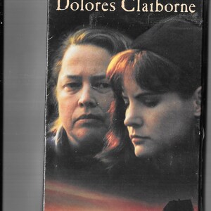May include: A VHS movie cover for "Dolores Claiborne" featuring Kathy Bates and Jennifer Jason Leigh. The cover art shows two women's faces, a silhouette of a person, and a house against a sunset.