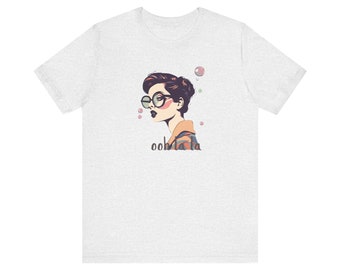 May include: A heather grey t-shirt with a graphic of a woman wearing round glasses and a colorful patterned shirt. The text "ooh la la" is printed below the graphic.