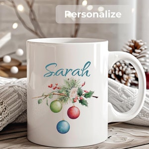 May include: White ceramic mug with a Christmas-themed design. The mug features a green pine branch with red berries, a white rose, and three ornaments in red, green, and blue. The name "Sarah" is written in blue cursive on the mug.