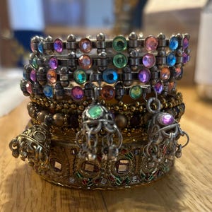 May include: A stack of four gold and silver bangles with colorful iridescent rhinestones and dangling charms. The bangles are decorated with intricate patterns and designs.
