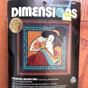 May include: A needlepoint kit titled "Oriental Moon Fan" featuring a framed design of a geisha with a fan. The artwork uses shades of red, orange, blue, and black. The frame size is 5" x 5" (13 x 13cm).