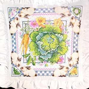 Puede incluir: Un cojín cuadrado bordado a punto de cruz con un borde blanco con volantes. El diseño presenta una col central rodeada de zanahorias, flores y conejos. El borde tiene un patrón de cuadros azules y blancos.