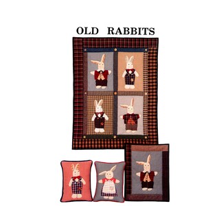 Könnte beinhalten: Eine Patchwork-Quiltdecke und dekorative Kissen mit applizierten Hasenfiguren in verschiedenen Outfits. Die Quiltdecke hat einen karierten Rand und den Text "OLD RABBITS" oben. Die Kissen haben ein ähnliches Design mit den Hasenfiguren in der Mitte.