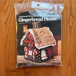 May include: A patchwork kit by Yours Truly for a Gingerbread House. The finished height is approx. 9" (22.9 cm). The house is made of fabric with red, white, and black accents, candy cane trim, and a heart-shaped design.