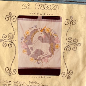 May include: A craft kit titled LA UNICORN featuring a white unicorn with a gold horn centered on a blue polka dot circle. The unicorn is surrounded by embroidered yellow and pink flowers. The kit includes 11 x 11 inch fabric and instructions.