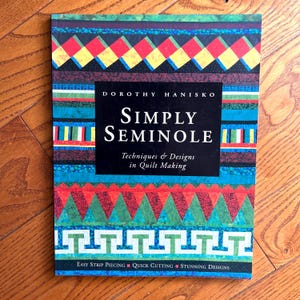 Könnte beinhalten: Ein Buch mit dem Titel "Simply Seminole" von Dorothy Hanisko, das Techniken und Designs für die Quiltherstellung vorstellt. Das Cover zeigt farbenfrohe geometrische Muster in Rot, Blau, Grün und Gelb. Das Buch erwähnt auch einfaches Streifen-Piecing, schnelles Schneiden und atemberaubende Designs.