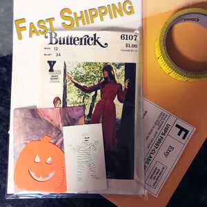 May include: A Butterick 6107 sewing pattern envelope featuring a Mary Quant design, accompanied by an orange pumpkin cutout, a white card with a mummy illustration, a roll of yellow measuring tape, and a shipping label on a tan envelope.