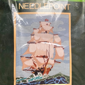 May include: Needlepoint kit featuring a detailed depiction of a tall ship sailing on a choppy sea. The ship has tan sails and a brown hull, set against a blue sky and water. The word "NEEDLEPOINT" is printed at the top.