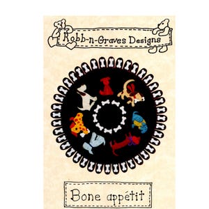 Può includere: Una toppa circolare in tessuto nero con un bordo di icone di ossa bianche e un anello centrale di ossa bianche. Sei illustrazioni di cani colorati sono disposte in cerchio. L'etichetta superiore recita Robb-n-Graves Designs e quella inferiore Bone appétit.