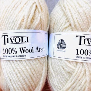 以下が含まれることがあります： オフホワイトのTivoli 100% Wool Aran毛糸玉2つ。各毛糸玉には、Tivoli、100% Wool Aran、Knits to Aran Patternsという黒い文字が印刷された白い紙ラベルが付いており、右側のラベルにはPure New Woolのロゴが記載されています。