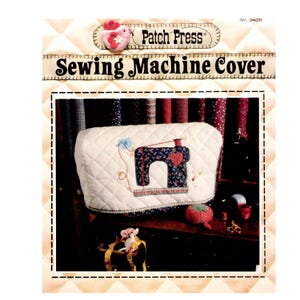 Può includere: Una copertura trapuntata color bianco sporco per macchina da cucire con un'applicazione di macchina da cucire floreale blu scuro e un cuore rosso. La copertura riporta il testo Patch Press Sewing Machine Cover e No. 3456. È visibile un puntaspilli rosso.