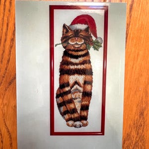 May include: A framed illustration of a tabby cat wearing a Santa hat and holding mistletoe. The cat has brown and orange stripes and is smiling. The frame is red, and the background is light green.