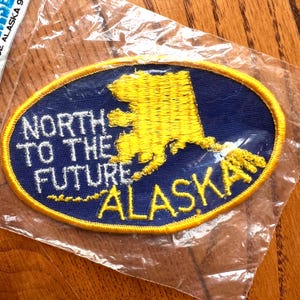 May include: An oval-shaped patch with a blue background and a gold embroidered outline. The patch features a gold embroidered outline of the state of Alaska and the text "NORTH TO THE FUTURE ALASKA" in white and gold.