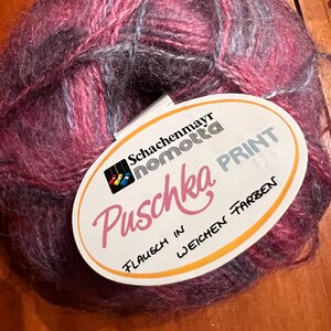 May include: A ball of Schachenmayr nomotta Puschka Print yarn in shades of pink, purple, and gray. The yarn has a soft, fluffy texture. A white label with the brand name and product details is attached.
