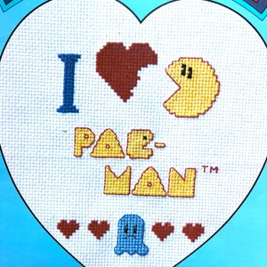 May include: A white heart-shaped cross-stitch with the text "I ♥ PAC-MAN™" in yellow and blue. There is a red heart, a blue ghost, and four red hearts at the bottom of the design.