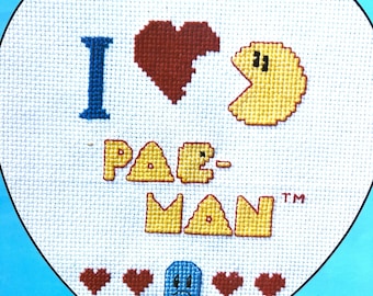 May include: A white heart-shaped cross-stitch with the text "I ♥ PAC-MAN™" in yellow and blue. There is a red heart, a blue ghost, and four red hearts at the bottom of the design.