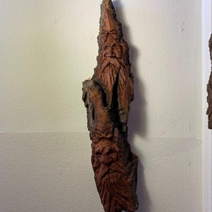 Op de afbeelding: Handgesneden houten sculptuur van een baardmansgezicht, aan de muur bevestigd. De sculptuur is gemaakt van een stuk natuurlijk hout, met ingewikkelde details. Het hout heeft een donkerbruine kleur.