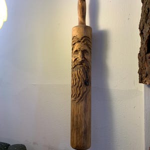 May include: A wooden rolling pin featuring a carved face of a bearded man. The rolling pin is crafted from light brown wood and has a handle at each end. The face is intricately carved, showcasing the man's hair, beard, and facial features.
