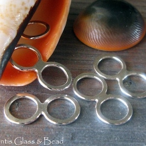 May include: Five silver-toned metal connector rings, each with two circles, are arranged on a wooden surface. The rings are designed to connect jewellery pieces together.