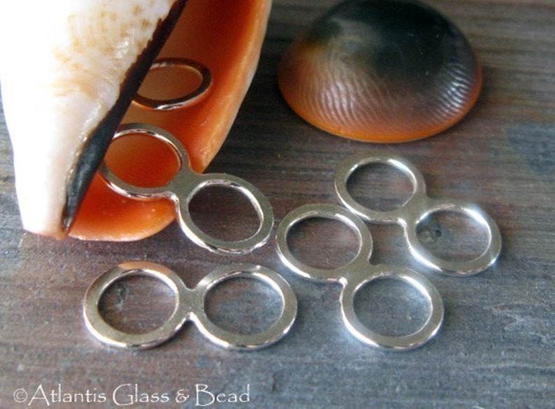 May include: Five silver-toned metal connector rings, each with two circles, are arranged on a wooden surface. The rings are designed to connect jewellery pieces together.