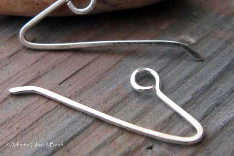 May include: Two silver earring hooks, one with a loop at the end and the other with a straight end. The hooks are on a wooden surface.
