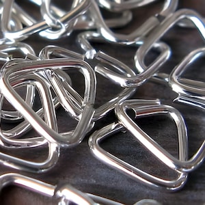 May include: Close-up of a pile of shiny, silver metal buckles. The buckles are triangular with rounded corners and a bar across one side. The objects are scattered across a dark surface.