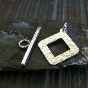 Op de afbeelding: Zilveren toggle-sluiting met een gehamerd vierkant ontwerp. De sluiting heeft een vierkante opening en een staaf met een ring aan het ene uiteinde.