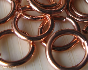 May include: A close-up of a group of copper jump rings. The rings are all the same size and are arranged in a random pattern. The rings are shiny and have a warm, metallic color.