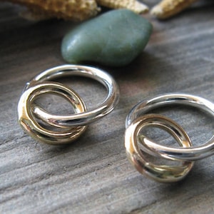 May include: Two interlocking rings, one silver and one gold, are shown on a wooden surface. The rings are smooth and have a polished finish.