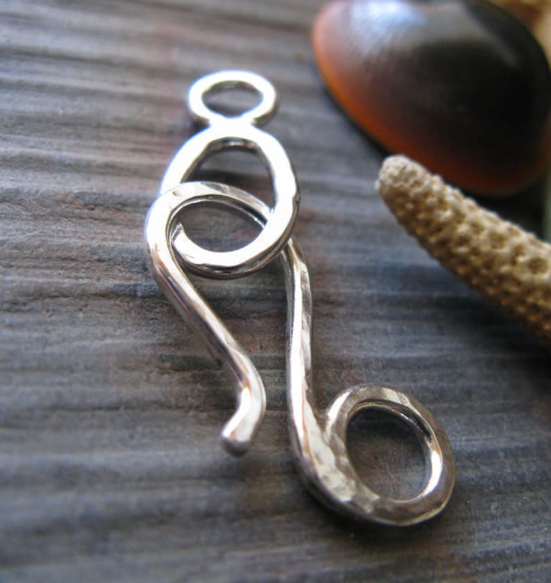May include: Silver hammered clasp with a unique design, featuring a loop and a hook. The clasp is made of polished silver and has a smooth, textured finish.
