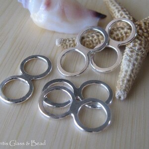 May include: Silver-toned metal double loop connectors. The connectors are in the shape of a figure eight with two circles. The connectors are arranged in a cluster on a wooden surface.