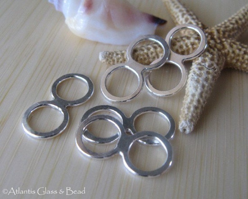 May include: Silver-toned metal double loop connectors. The connectors are in the shape of a figure eight with two circles. The connectors are arranged in a cluster on a wooden surface.