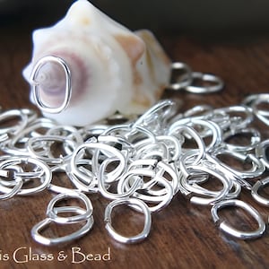 May include: Silver-coloured oval jump rings scattered on a wooden surface, with a seashell in the background. The open jump rings are for jewellery making or crafts. The text "Atlantis Glass & Bead" is visible.
