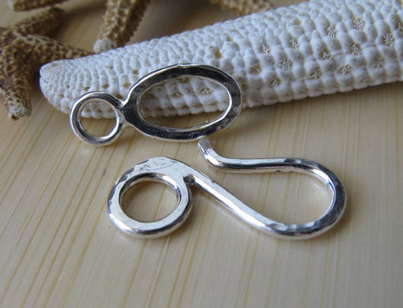 May include: Two silver-toned metal jewelry clasps with a hammered finish. The clasps are shaped like a circle with a loop on one side and a teardrop shape on the other.