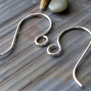 May include: Two silver earring hooks, one larger than the other, are shown on a wooden surface. The hooks are made of metal and have a smooth, shiny finish.