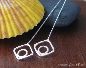 May include: Two silver hair pins with a square loop at the end. The loops have a smaller circle inside. The pins are on a wooden surface.
